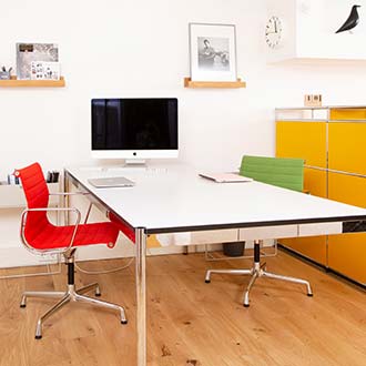 Shari Dietz' Bürozimmer mit weißem Schreibtisch, gelben Aktenschrank sowie Stühlen in intensivem Rot und Grün